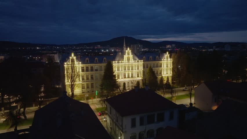 A wide aerial shot of the historic Liviu Rebreanu College in Bistrita, Romania. The grand building is beautifully illuminated against the twilight sky and city lights.