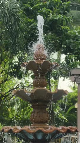 A vertical close-up shot of an ornate, three-tiered brown stone garden fountain with water gently cascading and bubbling amidst lush green tropical foliage and trees in soft daylight.
