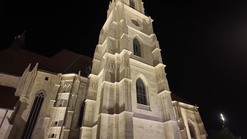 Evening walking tilt-up shot revealing the illuminated Gothic tower of St. Michael’s Church in Cluj-Napoca, Romania, emphasizing its detailed stonework and clock tower against the dark sky.