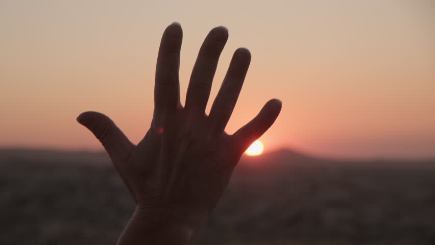Man holds his hand open, palm facing the sun, clenching it into fist at sunset in mountain valley