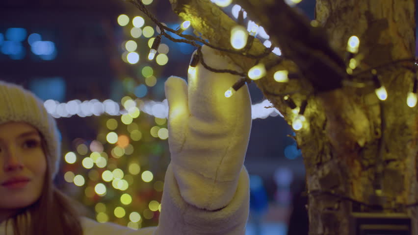 Close up of young woman in winter attire gently touching glowing festive lights while admiring them, warm golden glow enhances dreamy bokeh city background with blurred people passing by