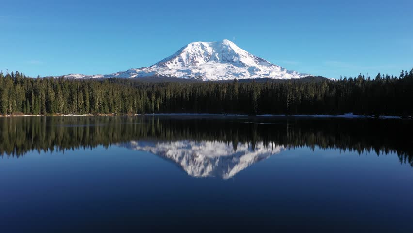 4K aerial drone footage of Takhlakh Lake and snow covered Mount Adams in Washington State USA during late fall early winter. Scenic reflection of mountain in calm water surrounded by evergreen forest 