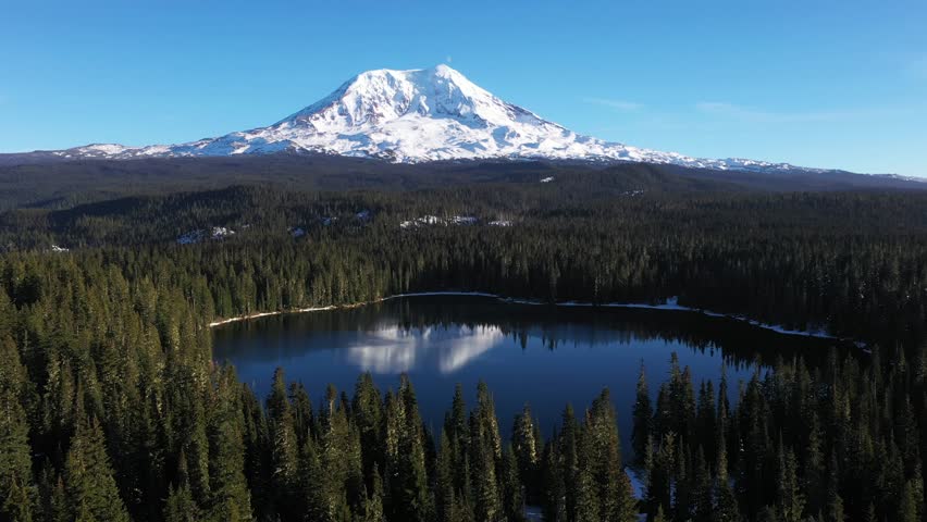 4K aerial drone footage of Takhlakh Lake and snow covered Mount Adams in Washington State USA during late fall early winter. Scenic reflection of mountain in calm water surrounded by evergreen forest 