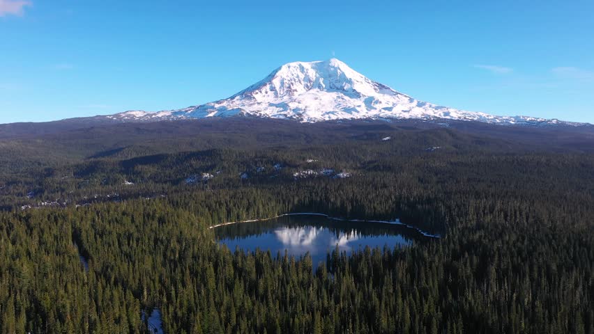 4K aerial drone footage of Takhlakh Lake and snow covered Mount Adams in Washington State USA during late fall early winter. Scenic reflection of mountain in calm water surrounded by evergreen forest 