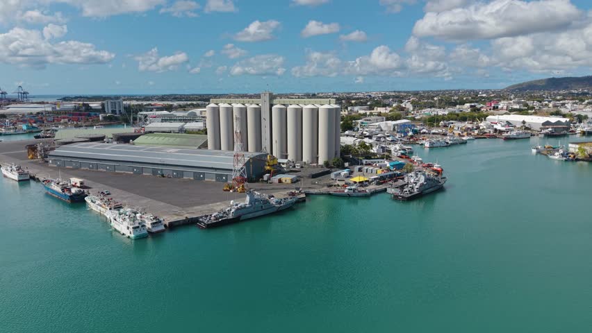 Aerial of the Port Louis harbor in Mauritius, featuring large industrial grain silos, warehouses, and docked ships against the backdrop of the city and blue water.