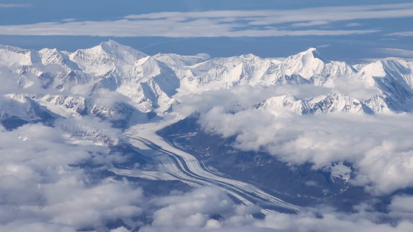 Aerial view of majestic snow-capped mountain peaks piercing through a sea of fluffy white clouds and a glacier, Denali, Alaska, United States.
