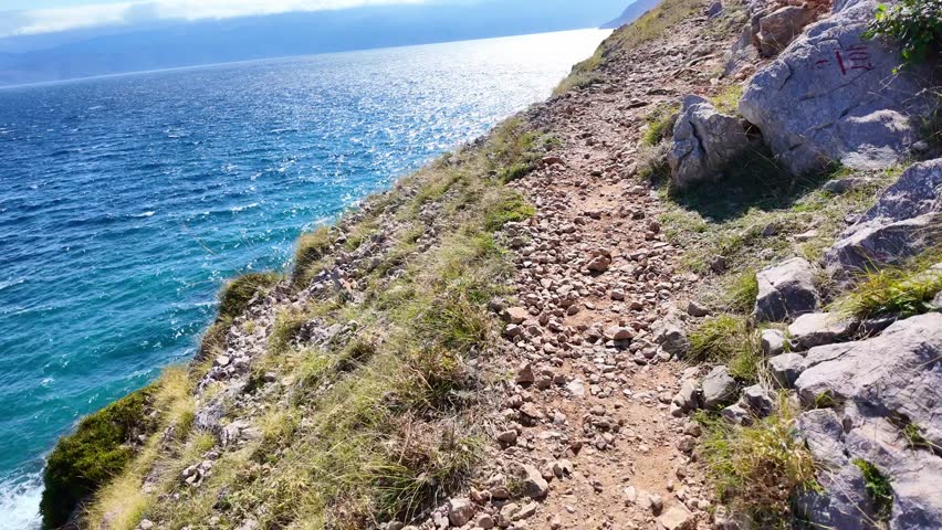 Coastal cliffside path with rugged terrain and vibrant blue sea under clear sky. Hiking trail near Baška, Krk island, Croatia.