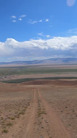 Vertical video of a car driving along a country road under blue sky. Altai desert and semi-desert environment. The village of Kokorya is visible in the valley.