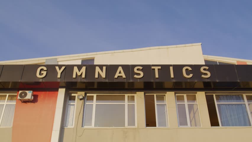 The exterior of an old gymnastics building glows under the warm sunset, showing classic Soviet-style architecture and faded signage.