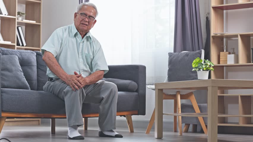 Asian elderly man knee pain sitting sofa holding knee living room with natural light and potted plant Elderly man sitting living room sofa holding knee pain gray hair glasses light shirt look