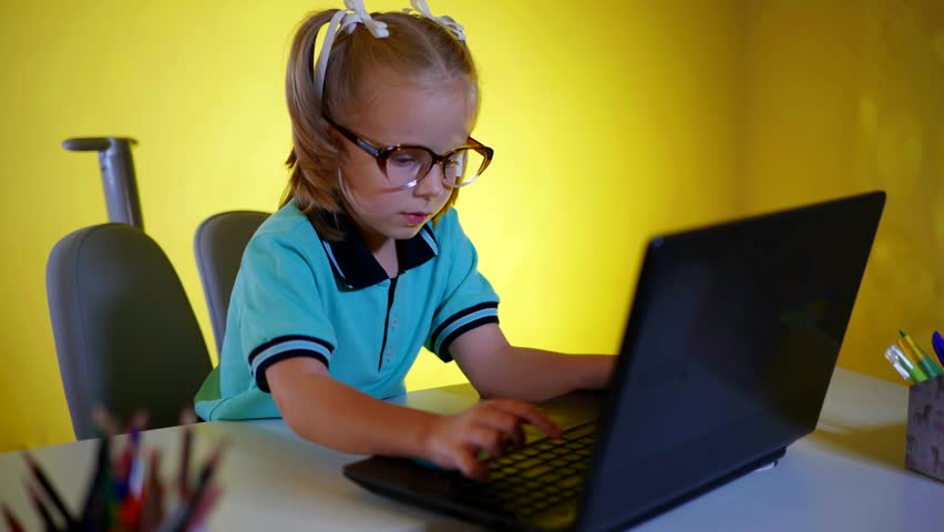Young girl in glasses intensely focused on typing on a laptop against a vibrant yellow background. Focused student with light hair tied back, using a notebook computer indoors.