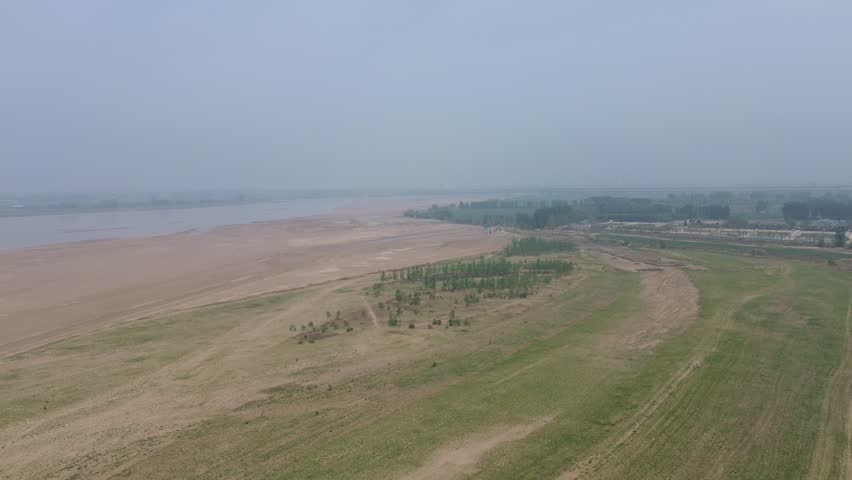 Aerial view with Zhengxin Yellow River Rail-Road Bridge in the distance, Zhengzhou, China, spring noon. Showcasing Yellow River sandbars, green fields, and misty scenery.