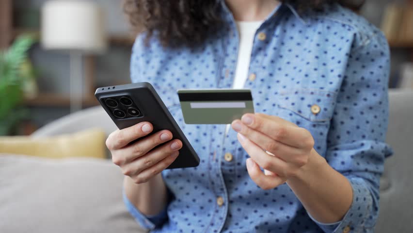 Close up of female hands using phone to make online transaction with credit card indoors at home. Shopper makes payment, pays bills, spends money on internet, e-commerce, e-shopping. Digital payment