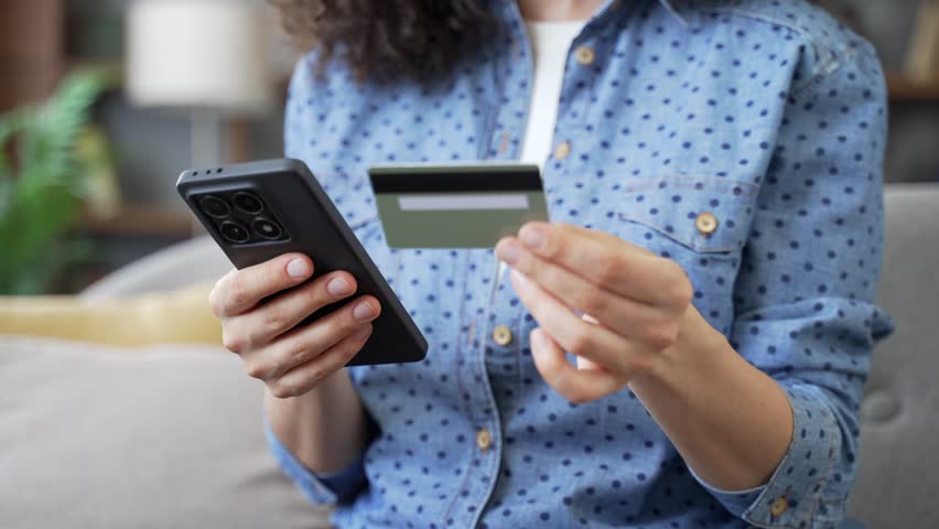 Close up of female hands using phone to make online transaction with credit card indoors at home. Shopper makes payment, pays bills, spends money on internet, e-commerce, e-shopping. Digital payment
