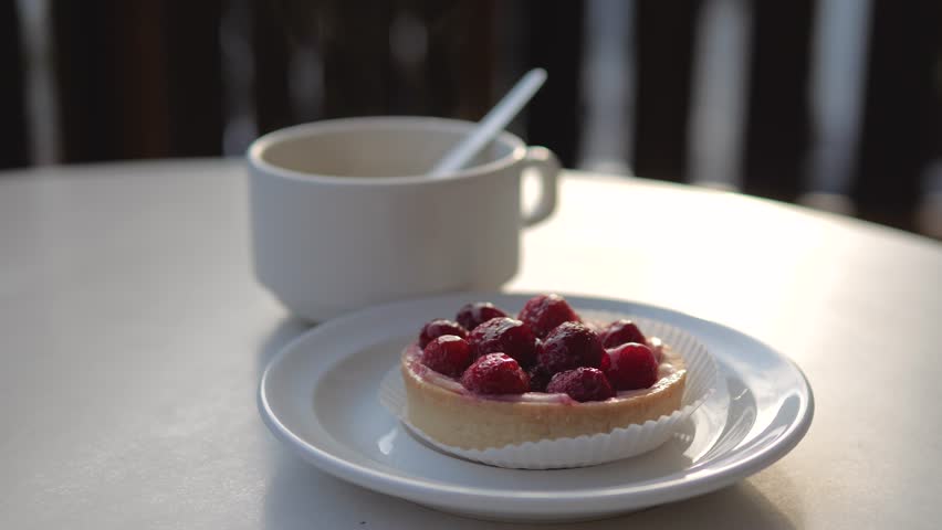 Morning sunlight illuminating delicious raspberry tartlet on white plate next to cup of steaming hot coffee on cafe table. Creating a cozy and inviting atmosphere for breakfast or a snack