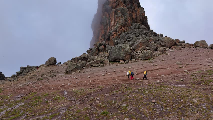 A group of hikers ascends the rocky slopes of Mount Kilimanjaro, Tanzania. Towering cliffs rise through the mist, creating a dramatic and rugged landscape