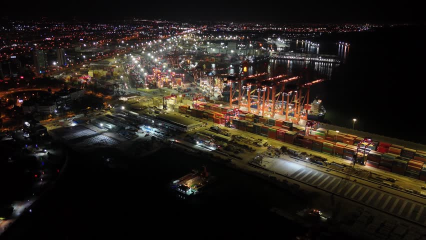 Nighttime aerial view of mersin port, turkey, with illuminated container terminals, cranes and cargo ships along the mediterranean coastline showing busy maritime trade operations