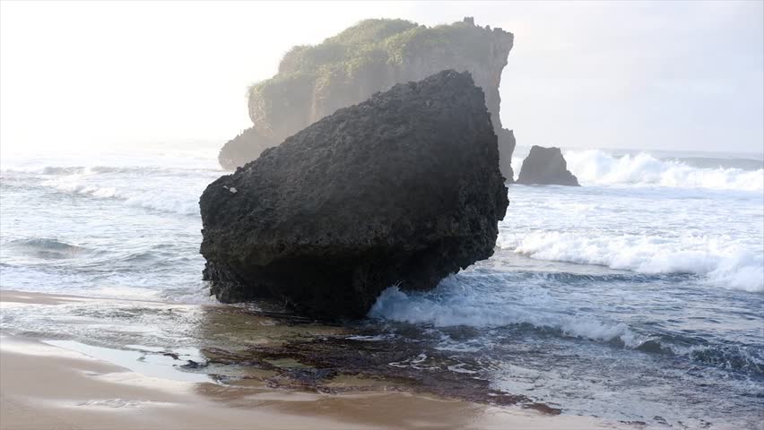 Large rock formation on sandy beach with ocean waves splashing at sunrise, natural coastal landscape with sea foam and misty atmosphere.