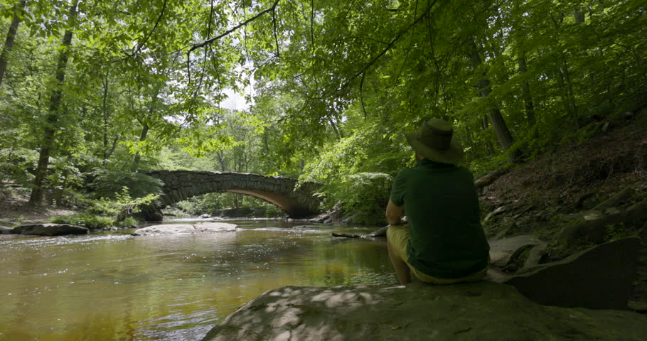 Boulder Bridge in Rock Creek Park, DC, is seen as a senior man in a wide-brim hat sits next to Rock Creek on a spring day. The wide shot tracks right to left in slow motion.
