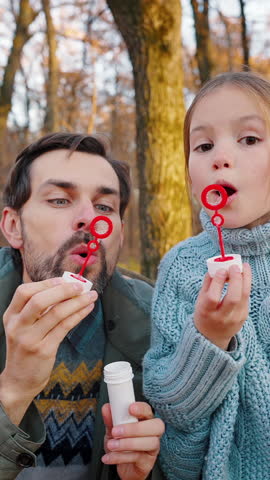 Young dad in casual clothes and little daughter blowing soap bubbles which flying on you. They smiling and talking. Sunny day in autumn park with yellow foliage. Happy family. Close up, slow motion