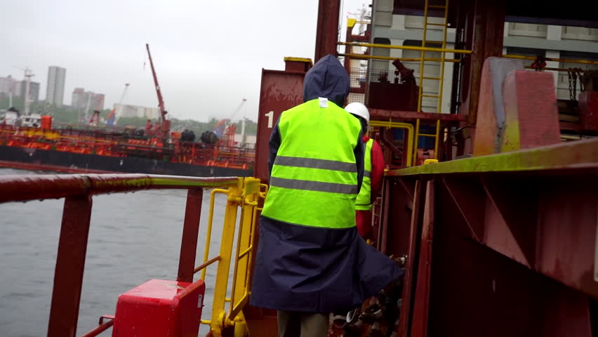 Sailor in bright reflective vest walks carefully along wet trembling deck. Jacket flashes against dark sea and steady rhythm of rain