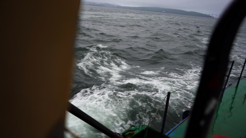 Waves crash rhythmically against ships hull spreading foam in air. Ship moves through turbulent water while spray glitters under pale light