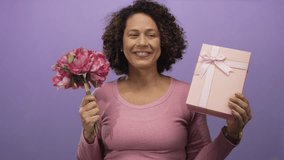 Woman smiling holds pink flower bouquet and gift box with both hands in purple studio; joy gratitude celebration surprise. - Powered by Shutterstock - Get 15% off with code: PIKWIZARD15