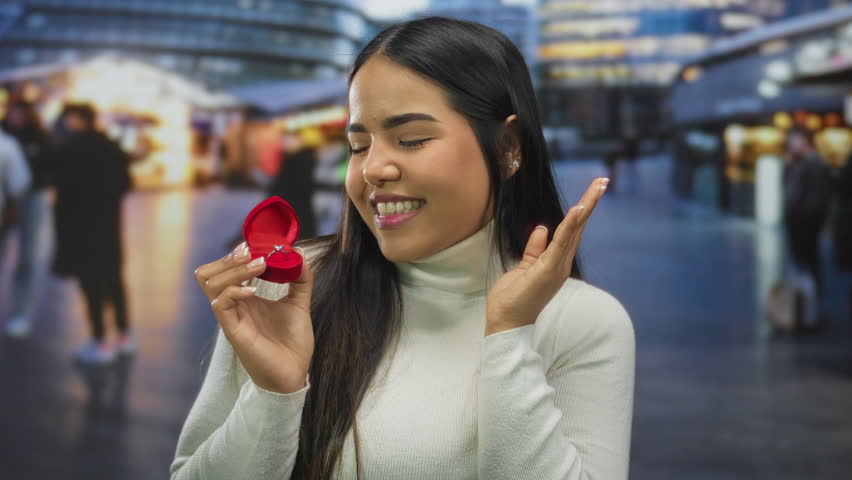 Woman smiling with engagement ring in red box on street evening lights city background facial expression.