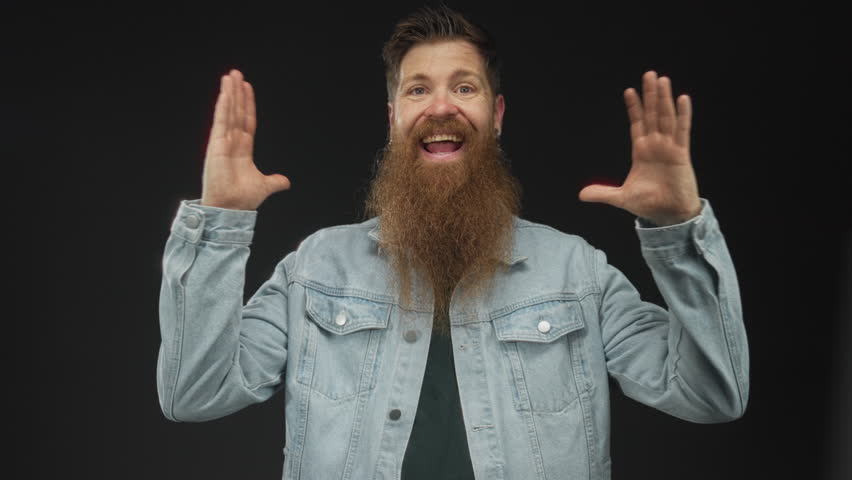 Man with beard wearing light denim jacket smiling broadly raising hands at eye level in studio; enthusiasm.