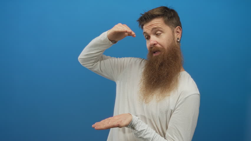 Man with long beard holds invisible object gesture with palms in blue studio; playfulness demonstration.