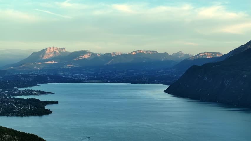 Aerial footage of a summer sunset above Lac du Bourget. The lake shines in evening light surrounded by mountains and forests. 