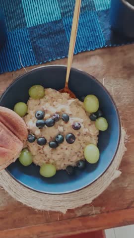 Cenital travel out shot of two bowls of oatmeal with blueberries and green grapes, accompanied by coffee and bread on a rustic wooden table with blue table mats.