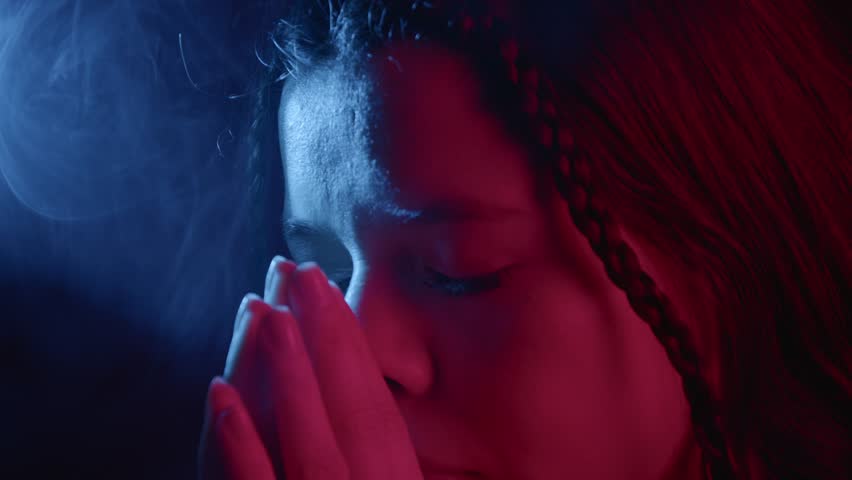 Close-up of a person in deep reflection, hands clasped, illuminated by dynamic red and blue lighting. Dramatic shadows enhance the contemplative mood.