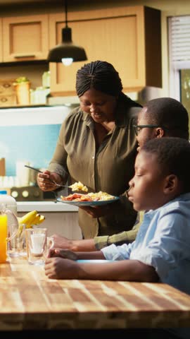 Vertical Video Parents and kids sitting together at breakfast table being served by the mother, serving omelette with bacon and goods. Cozy scene captures family bonding with joy for morning routine