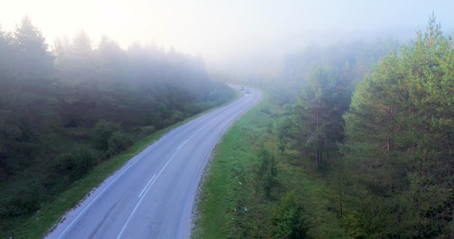 Aerial view of cars driving on a winding road through a misty green pine forest at sunrise