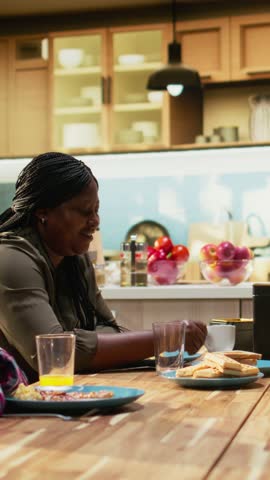 Vertical Video African American family sharing eggs and pastry for breakfast at home, parents serve omelettes, toast and fruit. Cozy kitchen moment captures warmth, relaxation and joyful togetherness