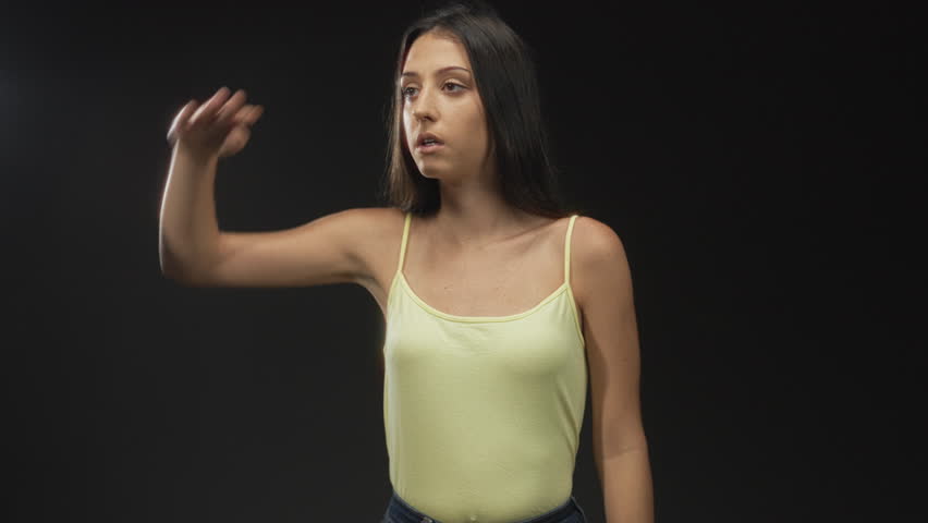 Woman shielding eyes with hand in studio against black backdrop, wearing yellow tank top and looking into the distance; curiosity.