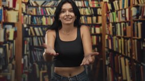 Young hispanic woman in black crop top offers open palms toward rows of bookshelves in library; excitement. - Powered by Shutterstock - Get 15% off with code: PIKWIZARD15