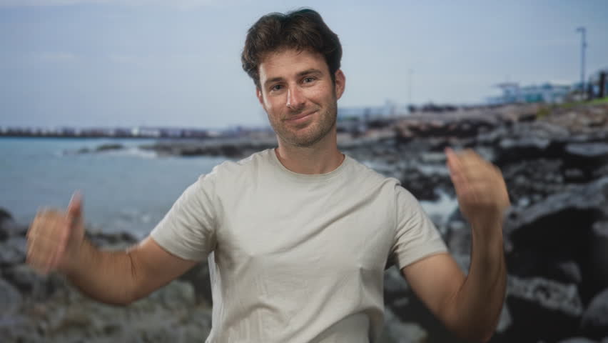 Man with bare arms forms heart with hands above head on rocky seaside street, smiling at camera; affection warmth.
