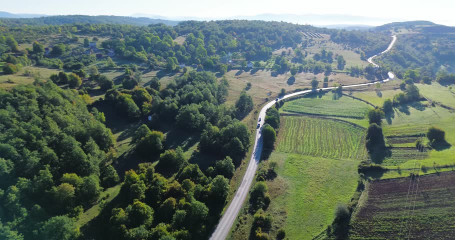 Cars traveling on a scenic rural road winding through lush green forests and agricultural fields