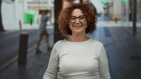 Woman smiling on city street in casual outfit with brunette curls and glasses waving outdoors during sunny day showing happy mood. - Powered by Shutterstock - Get 15% off with code: PIKWIZARD15