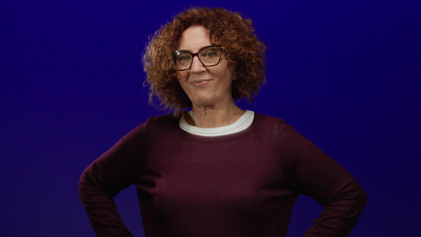 Woman with curly hair smiling confidently against an isolated blue background wearing glasses and casual attire, embodying positivity and friendliness in a studio setting.