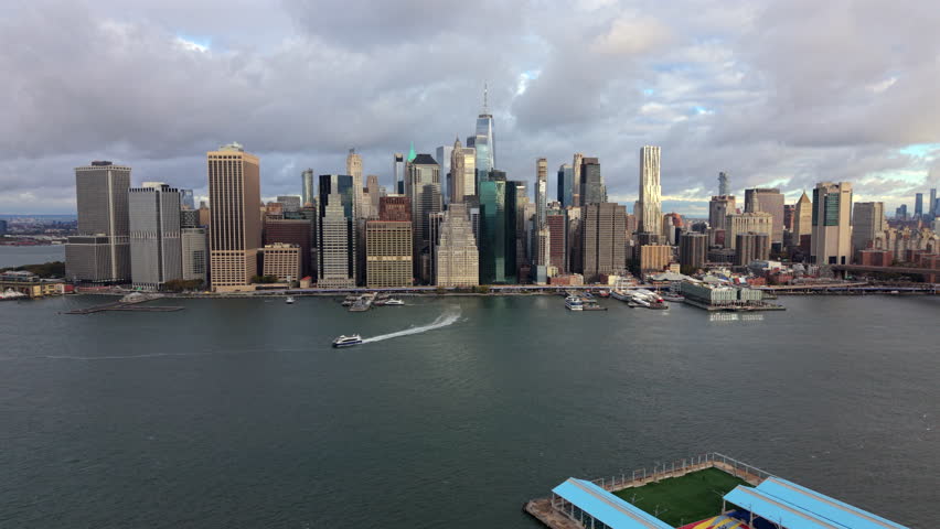 Aerial view of Lower Manhattan, iconic New York City skyline with ferry trail curving across the Hudson River, showcasing the contrast of urban density and open water under soft cloudy skies. New York