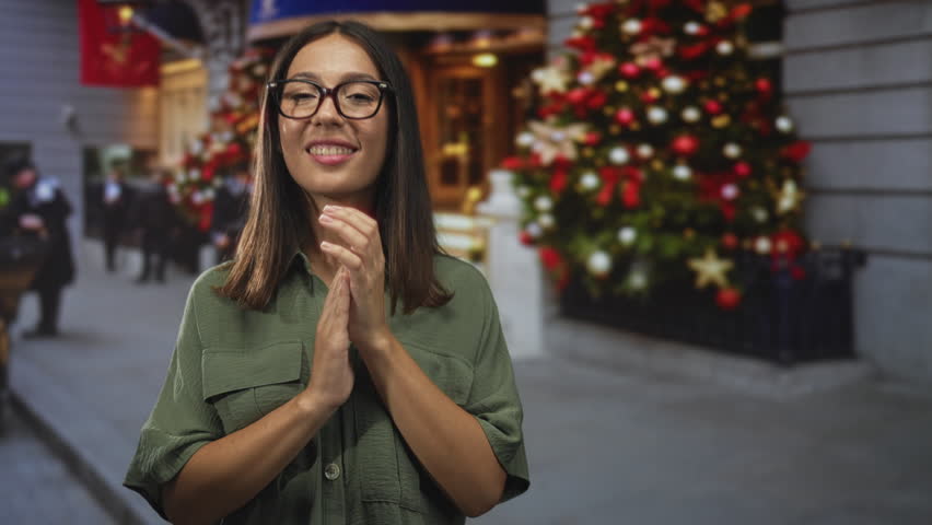 Woman clapping hands on city street in front of decorated building with lit christmas tree; holiday cheer.
