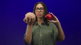 Young hispanic brunette woman holding model brain and red heart in studio on blue purple backdrop; conflict decision. - Powered by Shutterstock - Get 15% off with code: PIKWIZARD15