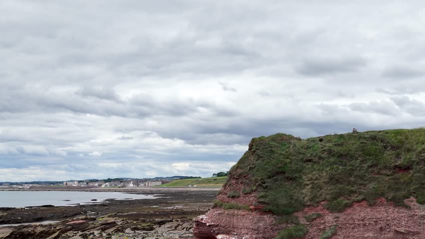 Camera pans right revealing Scottish red sandstone cliffs, eroded shoreline, grassy headland, and cloudy sky