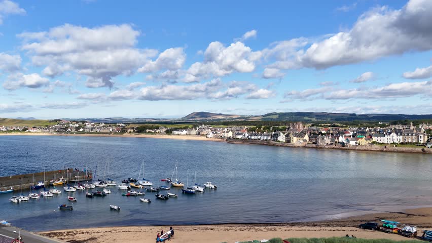 Drone pans over harbor with boats, waterfront village, and scenic landscape under bright daylight