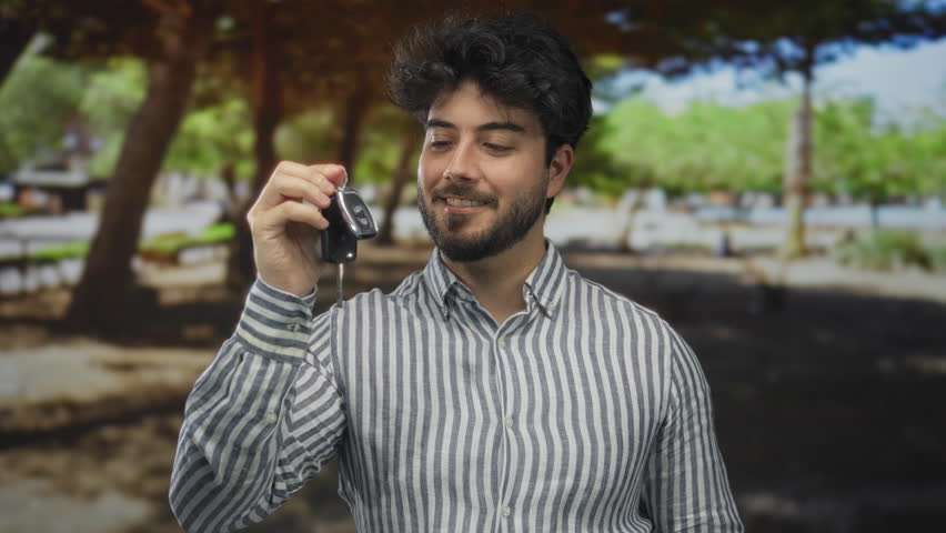 Young hispanic man with a beard holding car keys and a smartphone, standing in a shaded green park enjoying a sunny day.
