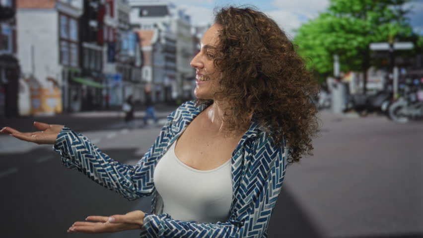 Woman smiling with open palms and bare chest visible on a city street in daylight, wearing white tank top and patterned shirt; friendly warmth joy.