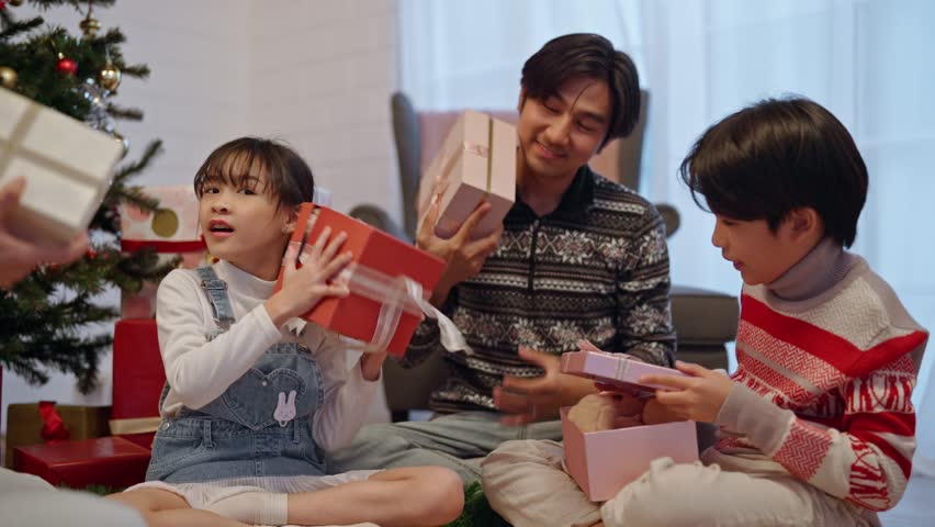 Asian young siblings opening present gift boxes under Christmas tree. Happy children boy and girl feeling joyful and excited with surprise gifts from family, celebrating festive Xmas together at home
